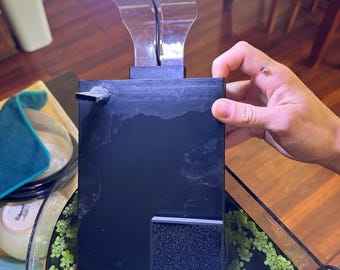 以下が含まれることがあります： 明るい白色LEDライトを備えた黒い水槽フィルター。フィルターは長方形で、小さな正方形のフィルターパッドが付いています。ライトはフレキシブルアームに取り付けられています。フィルターは、緑色の植物が入ったガラス水槽に配置されています。