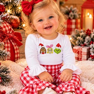 May include: A white long-sleeve shirt with ruffled cuffs and a graphic print of three characters wearing Santa hats, paired with red and white gingham pants featuring ruffled hems. The outfit includes white shoes and a red hair bow.