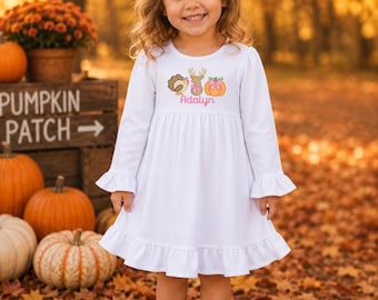 May include: A white long-sleeved dress with a ruffled hem and cuffs, featuring an embroidered design of a turkey, a deer, and a pumpkin with the name Adalyn in pink script. A white hair bow is worn with the dress and brown ankle boots.