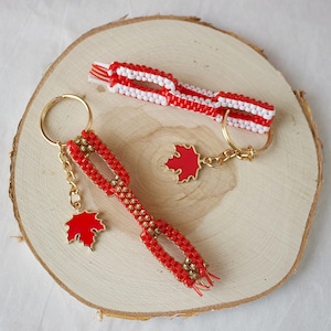 May include: Two keychains with red and white woven patterns and gold key rings. One keychain has a red maple leaf charm. The other keychain has a red maple leaf charm. The keychains are displayed on a wooden surface.
