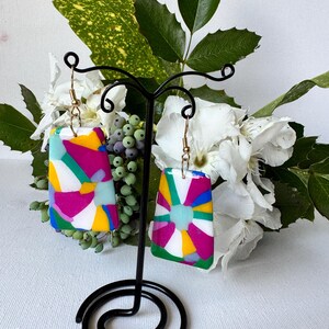Könnte beinhalten: Ein Paar rechteckige Ohrringe mit einem bunten Mosaikmuster in Rosa, Gelb, Blau, Grün und Weiß. Die Ohrringe hängen an goldfarbenen Haken und werden auf einem schwarzen Metallständer präsentiert. Im Hintergrund befinden sich weiße Blumen und grüne Blätter.