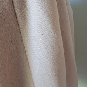 Może przedstawiać: Zbliżenie fragmentu jasnobeżowej tkaniny, prawdopodobnie płaszcza lub kurtki. Tkanina ma teksturowany splot, z widocznymi pionowymi fałdami i subtelnymi zmianami koloru. Obraz podkreśla fakturę i układ materiału.