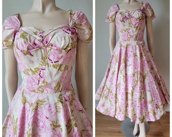 Może przedstawiać: Sukienka w stylu vintage z kwiatowym wzorem. Sukienka ma dekolt w kształcie serca, bufiaste rękawy i rozkloszowaną spódnicę. Tkanina jest biała z różowymi różami i zielonymi liśćmi. Sukienka jest prezentowana na manekinie.