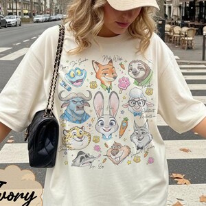 Pu&ograve; includere: T-shirt color avorio con una grafica colorata di personaggi di animali dei cartoni animati. La maglietta ha un girocollo e maniche corte. La parola "Ivory" &egrave; stampata in basso sulla maglietta.