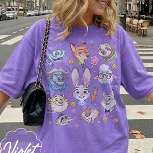 Pu&ograve; includere: T-shirt viola con grafica di personaggi di cartoni animati. La maglietta ha girocollo e maniche corte. La parola "Violet" &egrave; stampata in corsivo in basso a sinistra.