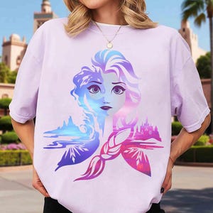 May include: A light purple t-shirt featuring a colorful graphic of a woman's face with blue and purple hair. The design includes a landscape and castle in shades of blue, pink, and purple.