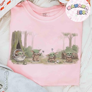 May include: A light pink t-shirt featuring a cartoon illustration of Grogu from The Mandalorian. The design shows Grogu in various poses in a forest setting. The shirt has the text "WANANA SHOP" in a circular logo.