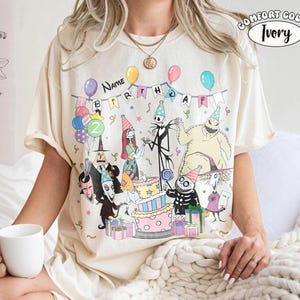 Op de afbeelding: Een ivoorkleurig t-shirt met een grafische print van personages uit The Nightmare Before Christmas op een verjaardagsfeestje. Het ontwerp bevat ballonnen, een taart met lagen, cadeautjes en een banner met de tekst Name BIRTHDAY. Comfort Color Ivory label is zichtbaar.