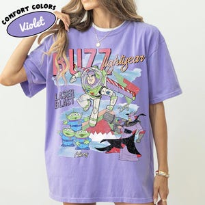 May include: Violet Comfort Colors t-shirt featuring a graphic of Buzz Lightyear, aliens, and Zurg from Toy Story. The design includes the text "Buzz Lightyear" and "Laser Blast". The shirt is a light purple color.