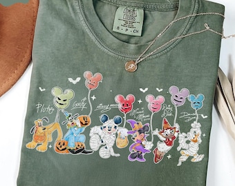 May include: Moss green Comfort Colors t-shirt featuring a Halloween-themed graphic of Disney characters. The design includes Pluto, Goofy, Mickey Mouse, Minnie Mouse, Daisy Duck, and Donald Duck, each holding a balloon. A silver necklace with a pendant is also visible.