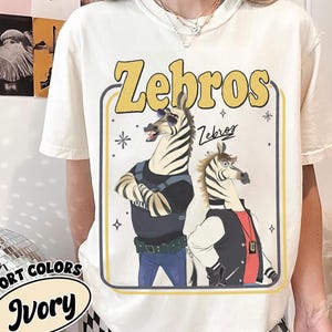 以下が含まれることがあります： 2匹のシマウマの漫画デザインが特徴のアイボリー色のTシャツ。「Zebros」の文字が黄色で描かれ、シマウマはサングラスとジャケットを着用しています。シャツの下部には「Fort Colors」と「Ivory」の文字も表示されています。