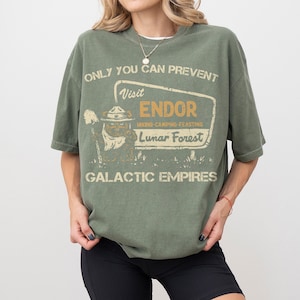 May include: An olive green t-shirt with the text "ONLY YOU CAN PREVENT" above a sign that reads "Visit ENDOR Lunar Forest." The shirt features a graphic of a bear-like creature in a ranger hat. The words "GALACTIC EMPIRES" are printed below the sign.