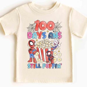 May include: Cream-colored t-shirt featuring a colorful graphic with the text "100 DAYS ANS STILL POPPIN". The design includes cartoon Spider-Man characters, popcorn, and spiderwebs. A fun and festive shirt for kids.