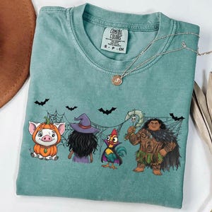 May include: A teal Comfort Colors t-shirt featuring a Halloween-themed graphic. The design includes cartoon characters of a pig in a pumpkin costume, a witch, a rooster, and a Polynesian warrior. Black bats and spiderwebs are also included.