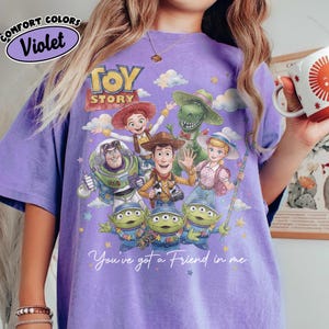 May include: Violet Comfort Colors t-shirt featuring a graphic of Toy Story characters. The design includes Woody, Buzz Lightyear, Jessie, Rex, Bo Peep, and the Little Green Men. The text "You've got a Friend in me" is also included.