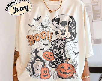 May include: Ivory-colored t-shirt with a Halloween-themed graphic. The design features Mickey Mouse in a skeleton costume, pumpkins, bats, spiderwebs, and the word "BOO!!". The shirt also has the words "Comfort Colors" and "Ivory" printed on it.
