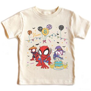 May include: Cream-colored t-shirt featuring cartoon Spider-Man characters. The design includes balloons, a birthday cake, and the number 5. The word "Name" is also visible. The shirt is decorated with bats and colorful confetti.