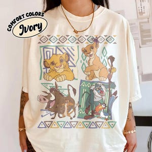 May include: Ivory-colored t-shirt featuring a colorful graphic of characters from The Lion King. The design includes Simba, Timon, Pumbaa, and Rafiki, with geometric patterns. The shirt has the words "Comfort Colors" and "Ivory" printed on it.