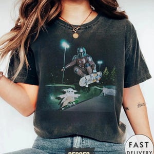 May include: Dark gray t-shirt featuring a graphic of The Mandalorian and Grogu skateboarding in a skate park. The design includes a detailed illustration of the characters and skateboards, set against a night scene with lights. The shirt has a vintage, worn look.