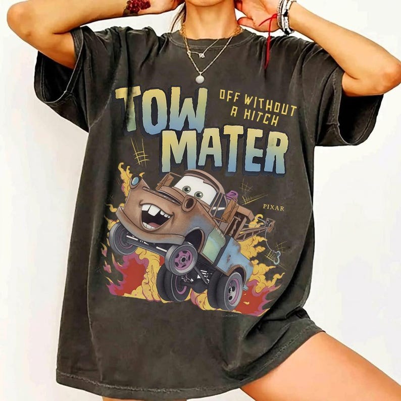 Może przedstawiać: Ciemnoszara koszulka z grafiką Złomka z filmu Pixar Cars. Projekt zawiera tekst "TOW MATER" i "OFF WITHOUT A HITCH" w kolorowej czcionce, z płomieniami i logo Pixar. Koszulka ma swobodny, obszerny styl.