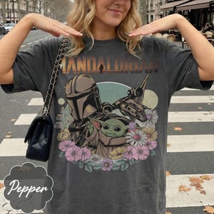 Op de afbeelding: Donkergrijs T-shirt met een afbeelding van The Mandalorian en Grogu, omringd door bloemen. De tekst "MANDALORIAN" staat bovenaan gedrukt. Het shirt heeft een vintage, versleten uitstraling.