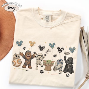 May include: An ivory-colored crew neck t-shirt featuring a graphic print of Star Wars characters including Chewbacca, a Stormtrooper, Grogu, R2-D2, Darth Vader, and BB-8 holding Mickey Mouse-shaped balloons, with small stars and lightsabers.
