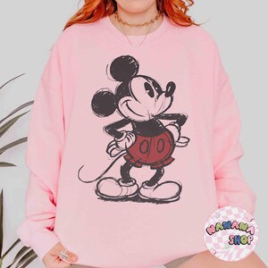 K&ouml;nnte beinhalten: Hellrosa Sweatshirt mit einem verblassten Mickey Mouse-Motiv in Schwarz, Rot und Wei&szlig;. Das Sweatshirt hat einen Rundhalsausschnitt und lange &Auml;rmel. Das Bild enth&auml;lt auch ein Logo mit der Aufschrift "MANANA SHOP".