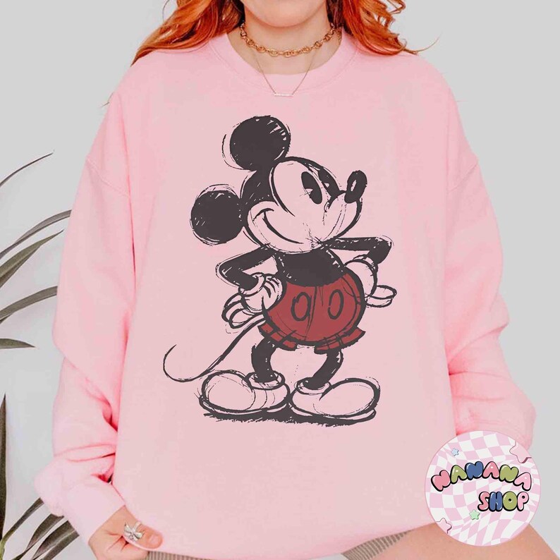 K&ouml;nnte beinhalten: Hellrosa Sweatshirt mit einem verblassten Mickey Mouse-Motiv in Schwarz, Rot und Wei&szlig;. Das Sweatshirt hat einen Rundhalsausschnitt und lange &Auml;rmel. Das Bild enth&auml;lt auch ein Logo mit der Aufschrift "MANANA SHOP".