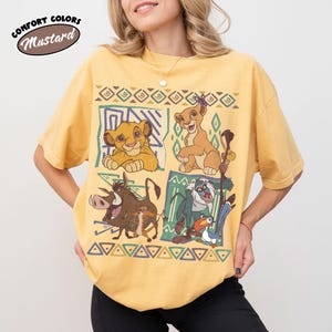 May include: Mustard-colored t-shirt featuring characters from The Lion King. The design includes Simba, Nala, Pumbaa, Timon, and Zazu, set against geometric patterns. The shirt has the words "Comfort Colors" and "Mustard" in a circular design.