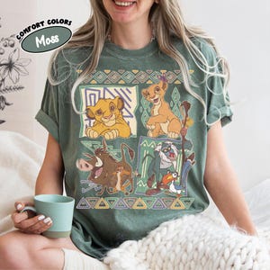 May include: Green t-shirt featuring characters from The Lion King. The design includes Simba, Timon, Pumbaa, and Rafiki, with geometric patterns. The shirt has the text "Comfort Colors" and "Moss" in a circular design.