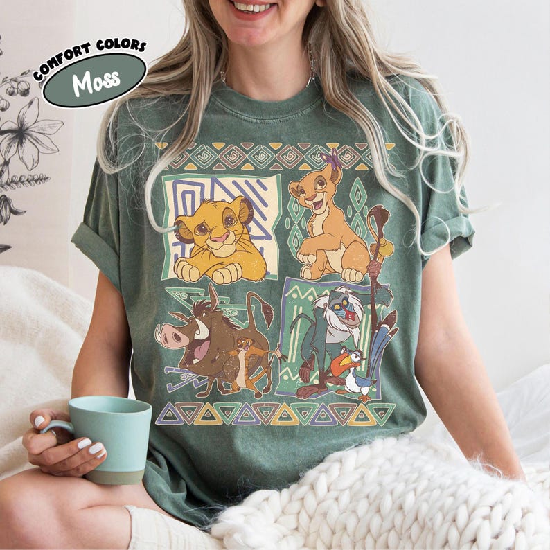 May include: Green t-shirt featuring characters from The Lion King. The design includes Simba, Timon, Pumbaa, and Rafiki, with geometric patterns. The shirt has the text "Comfort Colors" and "Moss" in a circular design.