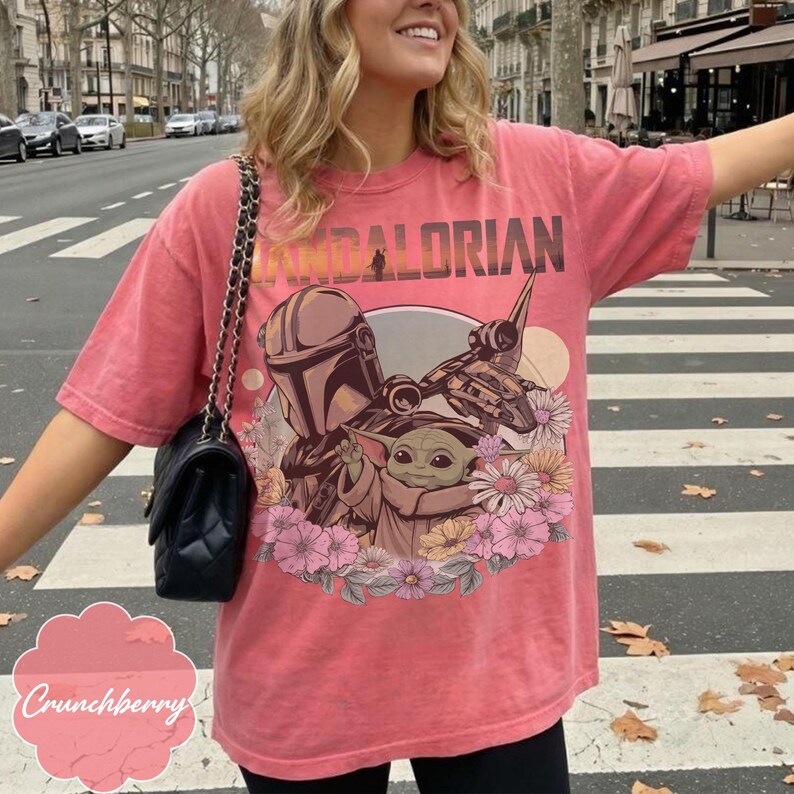 Op de afbeelding: Koraalrood T-shirt met een afbeelding van The Mandalorian en Grogu, omringd door bloemen. Het woord "MANDALORIAN" staat in vintage stijl bovenaan gedrukt. Het shirt heeft een relaxte pasvorm.