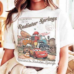 Op de afbeelding: Wit T-shirt met een kleurrijke afbeelding van personages uit de film Cars, waaronder Lightning McQueen. Het ontwerp bevat de tekst "California Est. 2012 Radiator Springs" en "Welcome to Radiator Springs."