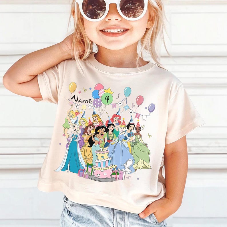 May include: A beige t-shirt featuring a colorful graphic of Disney princesses celebrating a birthday. The design includes a cake, balloons, and the number 4. The word "Name" is also visible.