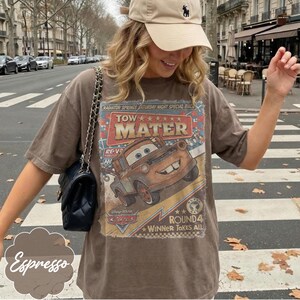 May include: Espresso-colored t-shirt featuring a graphic of Tow Mater from the movie Cars. The design includes the text "TOW MATER" and "RADIATOR SPRINGS SATURDAY NIGHT SPECIAL RACE". The shirt is worn with a beige cap and a black purse.