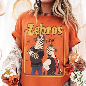 以下が含まれることがあります： オレンジ色のTシャツ。黄色で「Zebros」の文字、2匹の漫画のシマウマが描かれています。1匹は警察の制服、もう1匹はジャケットを着ています。シャツの袖は折り返されています。