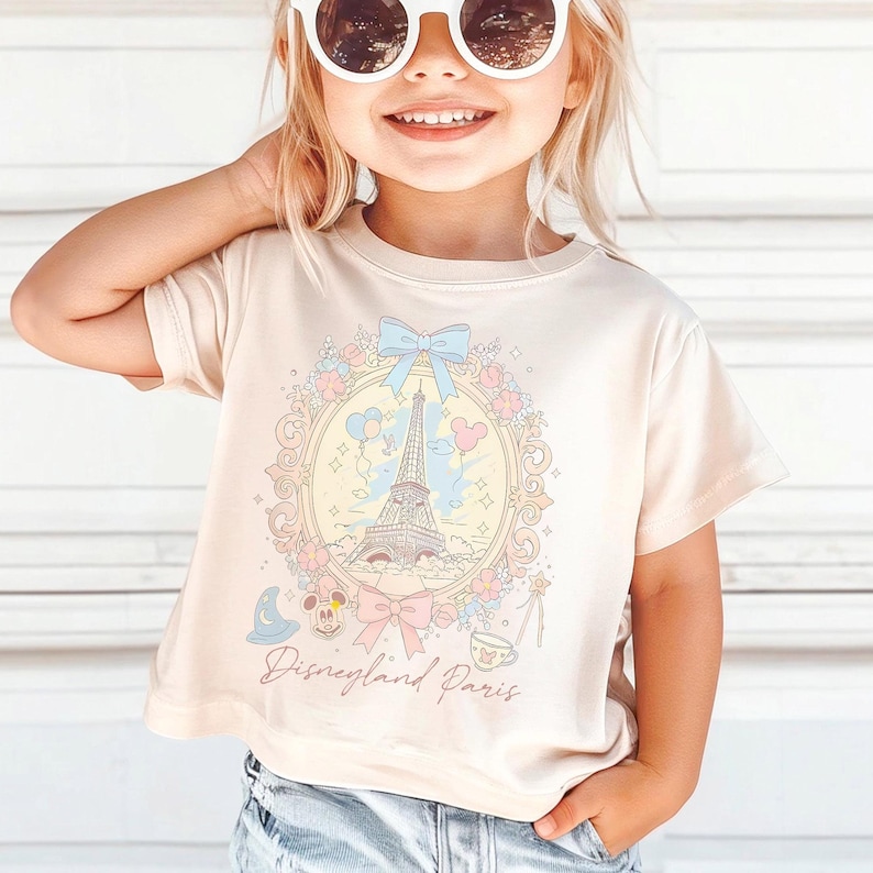 Peut inclure: Un t-shirt beige clair avec une illustration pastel de la Tour Eiffel, des ballons et des motifs floraux dans un cadre d&eacute;coratif. Le texte "Disneyland Paris" est imprim&eacute; en dessous. Le t-shirt est port&eacute; par un enfant.