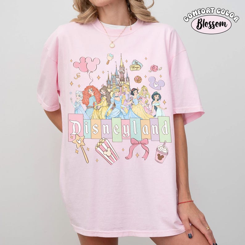 May include: Pink t-shirt with a Disneyland graphic featuring cartoon princesses, a castle, and other themed icons. The shirt has the words "Disneyland" in colorful letters and the words "Comfort Color Blossom" in a circle.