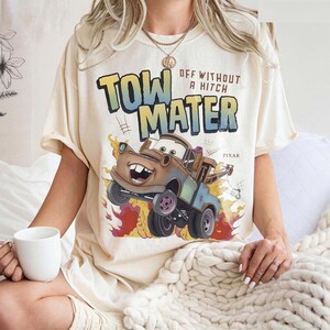 Może przedstawiać: Kremowa koszulka z grafiką Złomka z filmu Pixar Auta. Projekt zawiera napis "TOW MATER" w kolorze niebieskim i ż&oacute;łtym, z napisem "OFF WITHOUT A HITCH" powyżej. Obraz przedstawia Złomka z płomieniami.
