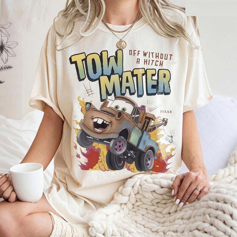 Może przedstawiać: Kremowa koszulka z grafiką Złomka z filmu Pixar Auta. Projekt zawiera napis "TOW MATER" w kolorze niebieskim i ż&oacute;łtym, z napisem "OFF WITHOUT A HITCH" powyżej. Obraz przedstawia Złomka z płomieniami.