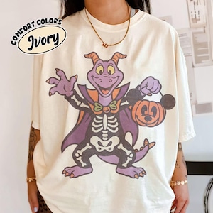 May include: An ivory t-shirt featuring a graphic of a purple dragon character wearing a skeleton costume and a cape, holding an orange pumpkin bucket with a Mickey Mouse ear silhouette. The text COMFORT COLORS IVORY appears in the top left corner.