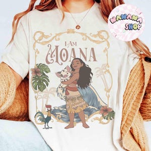 May include: Cream-coloured t-shirt with a graphic of Moana holding a pig, and the text "I AM MOANA" in a decorative frame. The design includes tropical elements like flowers, palm trees, and a rooster. The shirt has a round neck.