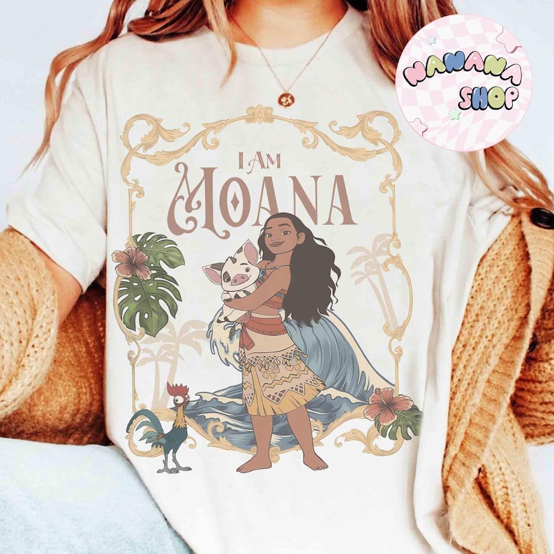 May include: Cream-coloured t-shirt with a graphic of Moana holding a pig, and the text "I AM MOANA" in a decorative frame. The design includes tropical elements like flowers, palm trees, and a rooster. The shirt has a round neck.