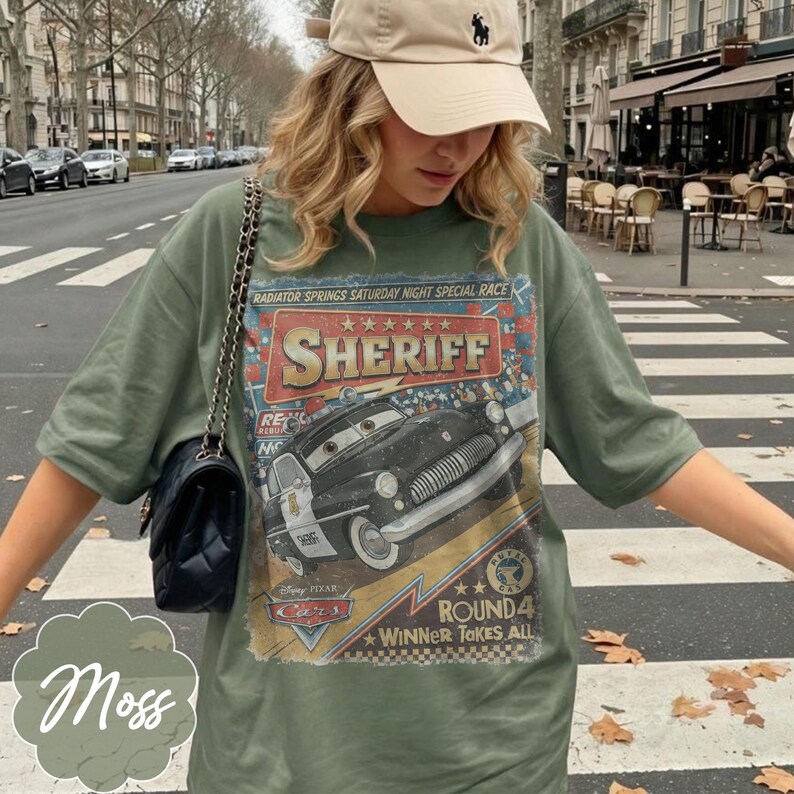 May include: Olive green t-shirt featuring a graphic of a black and white sheriff car from the movie Cars. The shirt has the text "SHERIFF" and "RADIATOR SPRINGS SATURDAY NIGHT SPECIAL RACE".