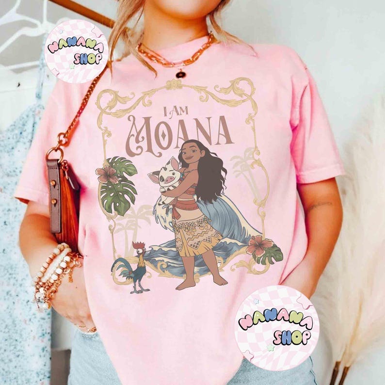 May include: Pale pink t-shirt with a graphic of Moana and a pig, with the text "I AM MOANA" in a decorative frame. The design includes tropical flowers and leaves. The shirt has short sleeves.