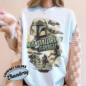 May include: Light blue t-shirt featuring a graphic from the Star Wars series, "The Mandalorian and Grogu." The design includes images of the Mandalorian, Grogu, and other characters, with the phrase "This is the Way."