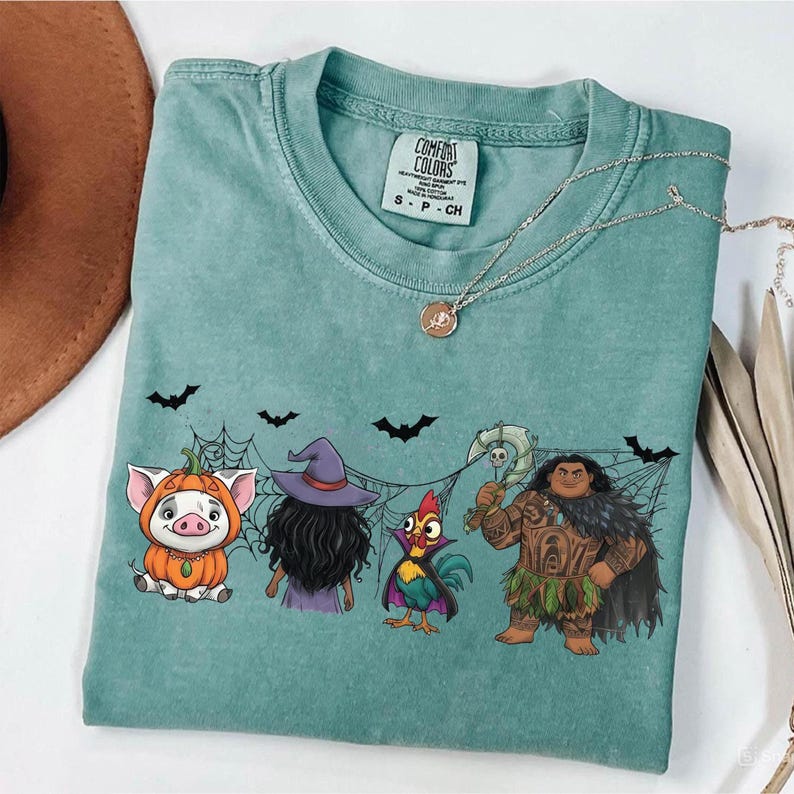 May include: A teal Comfort Colors t-shirt featuring a Halloween-themed graphic. The design includes cartoon characters of a pig in a pumpkin costume, a witch, a rooster, and a Polynesian warrior. Black bats and spiderwebs are also included.