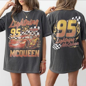 Puede incluir: Camiseta gris oscuro con Lightning McQueen de la pel&iacute;cula Cars. El gr&aacute;fico frontal incluye el n&uacute;mero 95, una bandera a cuadros y el nombre "McQueen". La parte trasera muestra "95" con llamas y "Lightning McQueen."
