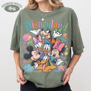 K&ouml;nnte beinhalten: Ein olivgr&uuml;nes T-Shirt mit einer bunten Grafik von Disney-Figuren, darunter Mickey Maus, Minnie Maus, Donald Duck, Daisy Duck, Goofy und Pluto. Der Schriftzug Disneyland steht oben, erg&auml;nzt durch die W&ouml;rter yay, whoa, magic und fun.