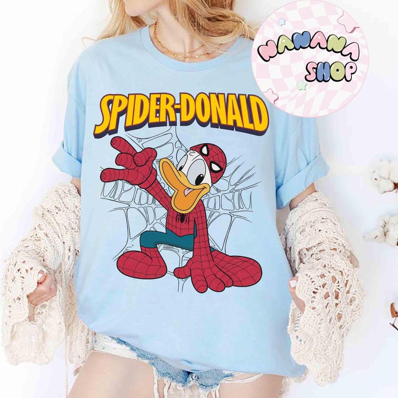 May include: Light blue t-shirt featuring a cartoon illustration of Donald Duck as Spider-Man, with the text "SPIDER-DONALD" in yellow. The design includes a web background and the character in a Spider-Man suit.